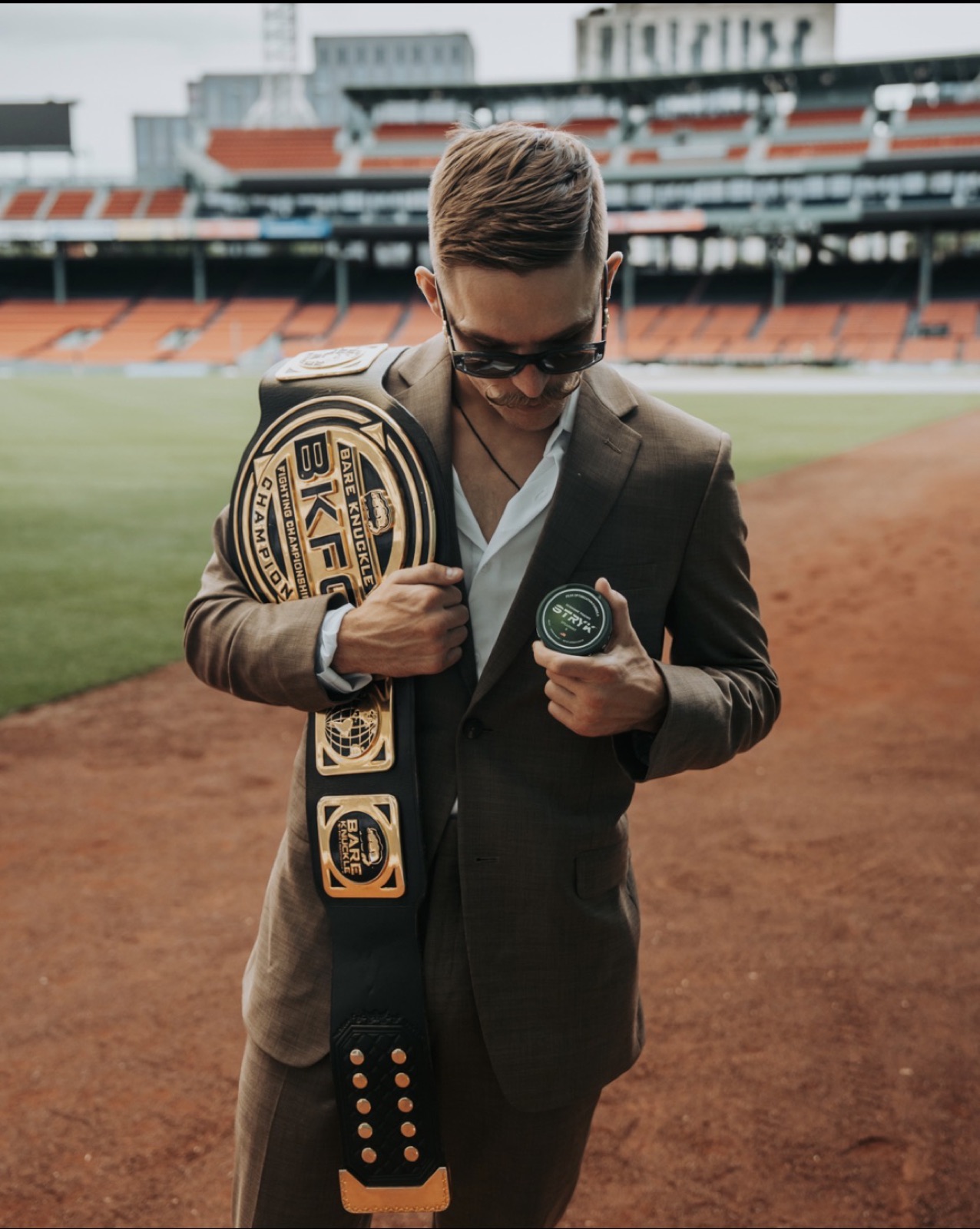 Kai Stewart in a suit with the BKFC featherweight belt on the field at Fenway Park
